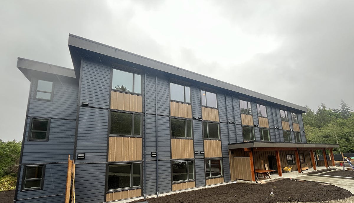 A modern three-story building with a dark gray exterior, wooden panel accents, and large windows, set against a backdrop of trees and a cloudy sky.