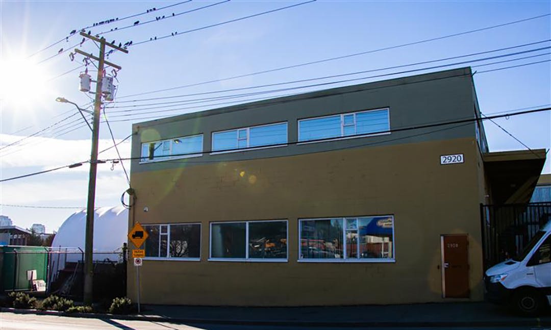 A two-story industrial building on a sunny day with power lines overhead, birds on wires, and a parked van at the right.