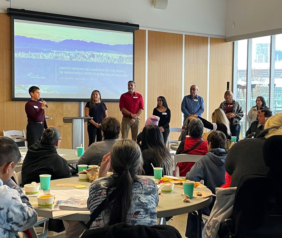 Indigenous City staff standing at the front of the room with Indigenous attendees sitting around tables