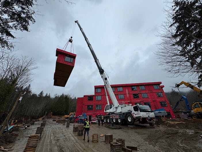 A large crane lifts a red modular building section at a construction site, where a multi-story red structure is being assembled amid trees, construction equipment, and workers coordinating the operation on the ground.