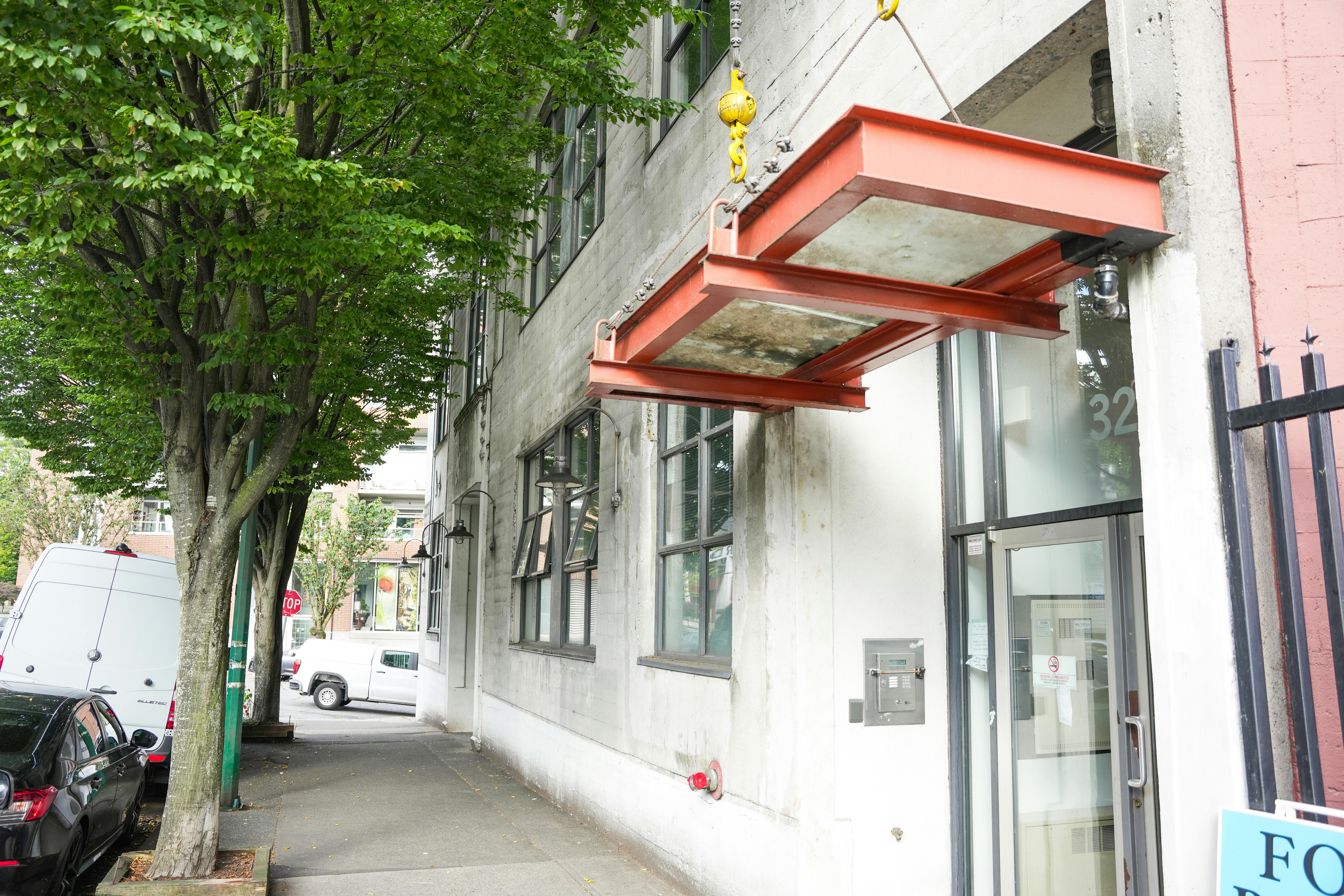 A tree-lined sidewalk in front of an industrial-style building at 321 Railway Street with large windows and a suspended red metal awning. 
