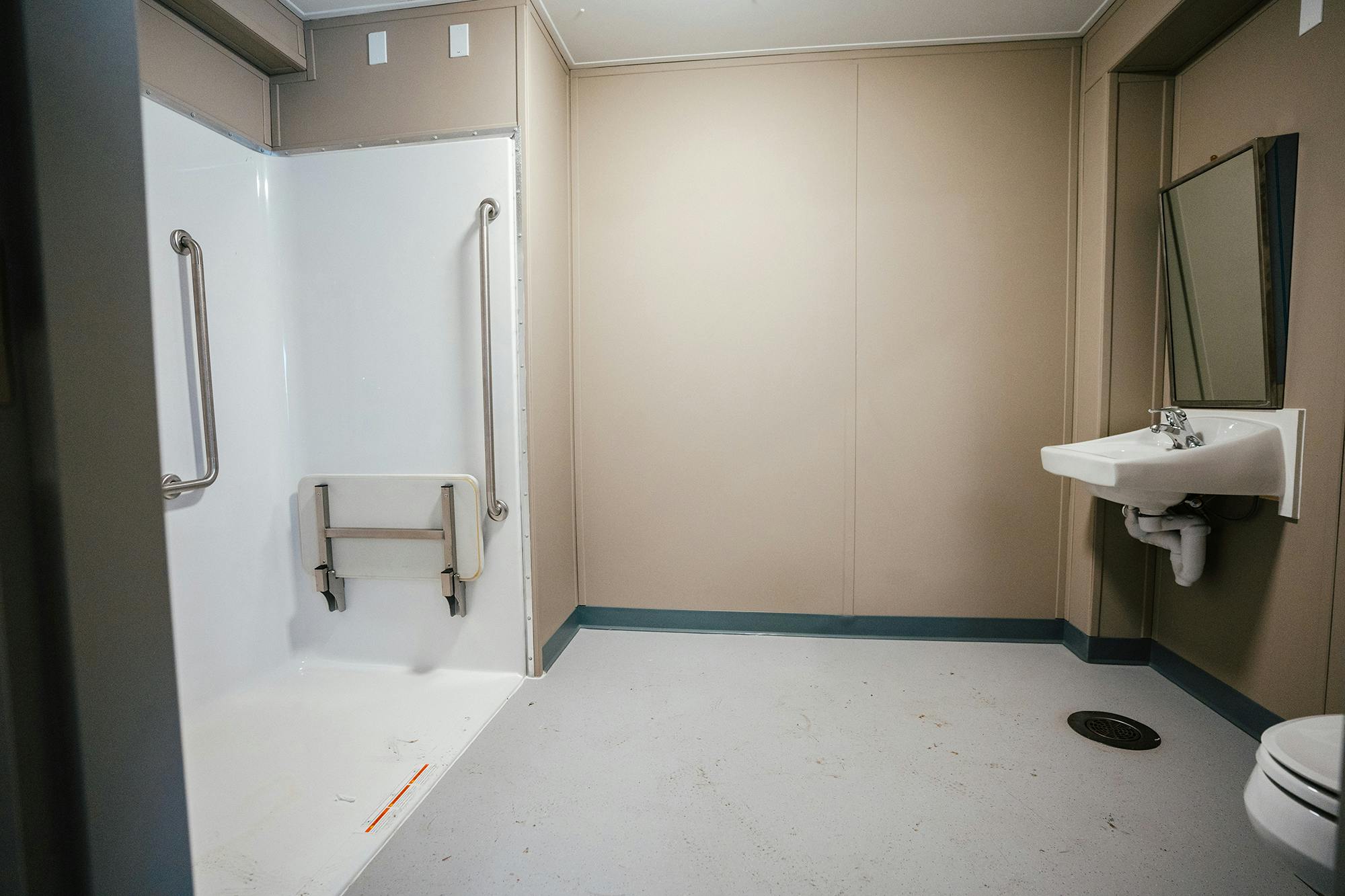 A spacious, accessible bathroom with a roll-in shower featuring grab bars and a foldable shower seat, along with a sink and a mirror on the opposite wall.