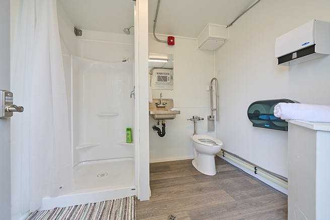Interior of a modular accessible washroom featuring a shower with a curtain, a toilet with a grab bar, a wall-mounted sink with a mirror above, and dispensers for paper towels and toilet paper.