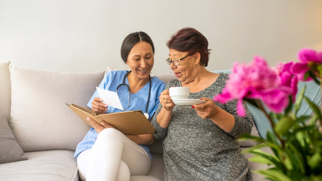 Clinician holding a folder with a paper smiling and a resident holding tea, talking to each other on a couch