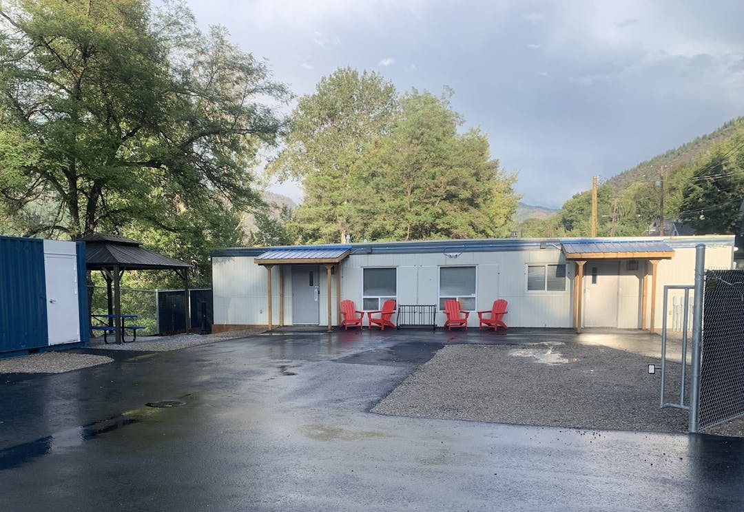 Outdoor scene with small buildings, wet asphalt, red chairs, green trees, and a mountain partially obscured by clouds.