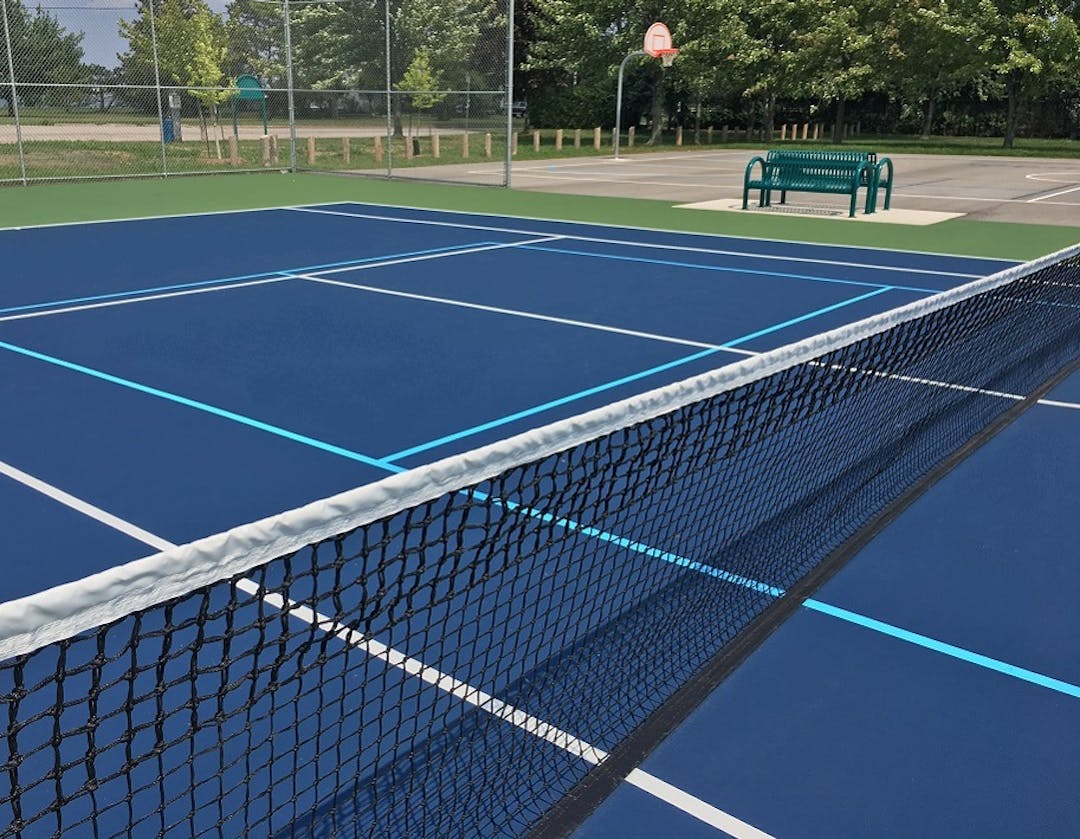 Riverview Neighbourhood Park tennis court with new blue playing surface, net, benches and basketball court in distance.